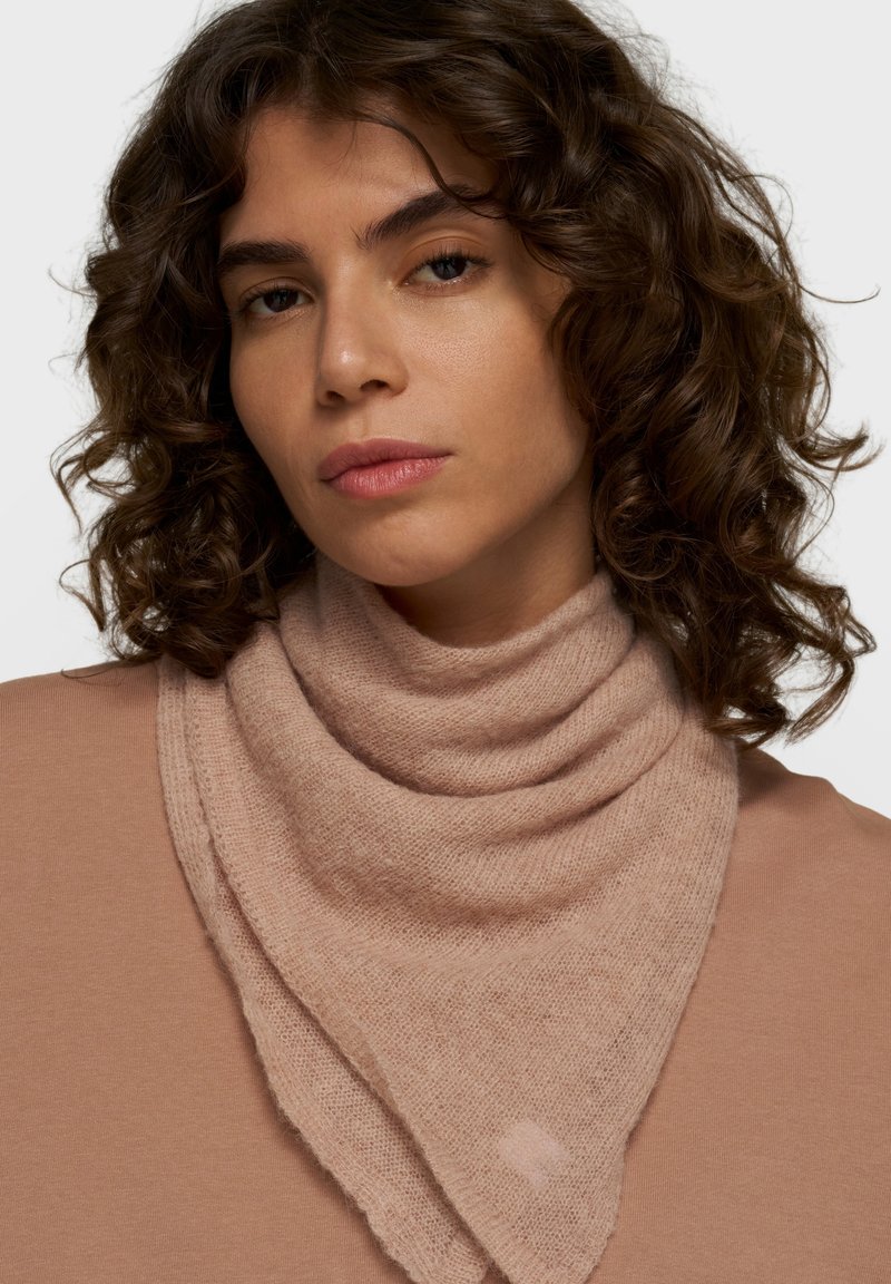 Mujer con el pelo corto, rizado y castaño lleva un jersey marrón claro y una bufanda de punto texturizada a juego, de frente sobre un fondo liso.