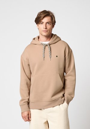 Jeune homme aux cheveux bruns portant un sweat à capuche beige avec une poche avant et des cordons à motif, associé à un pantalon de couleur claire.