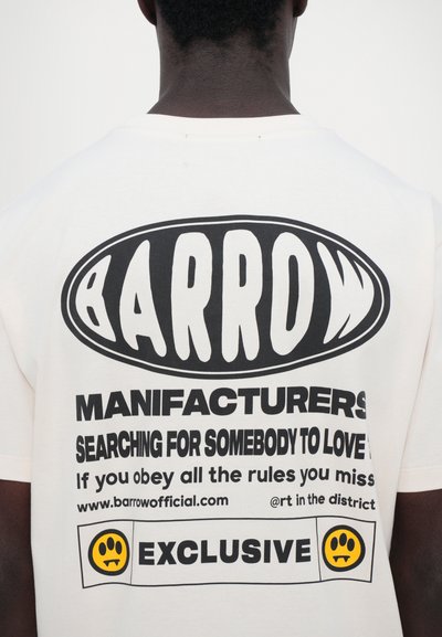T-shirt en coton crème avec un texte noir audacieux : « BARROW MANUFACTURERS », accompagné de graphiques et d'un logo avec un visage souriant. Coupe décontractée, manches courtes.
