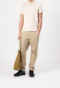 Chemise polo beige clair pour homme à manches courtes, pantalon kaki et baskets noires. Tiens un sac fourre-tout de couleur moutarde avec une bandoulière.