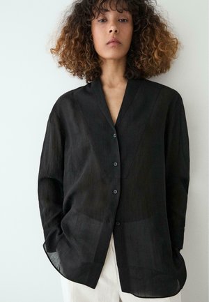Personne aux cheveux bouclés portant une chemise noire ample boutonnée et un pantalon blanc sur un fond clair uni.