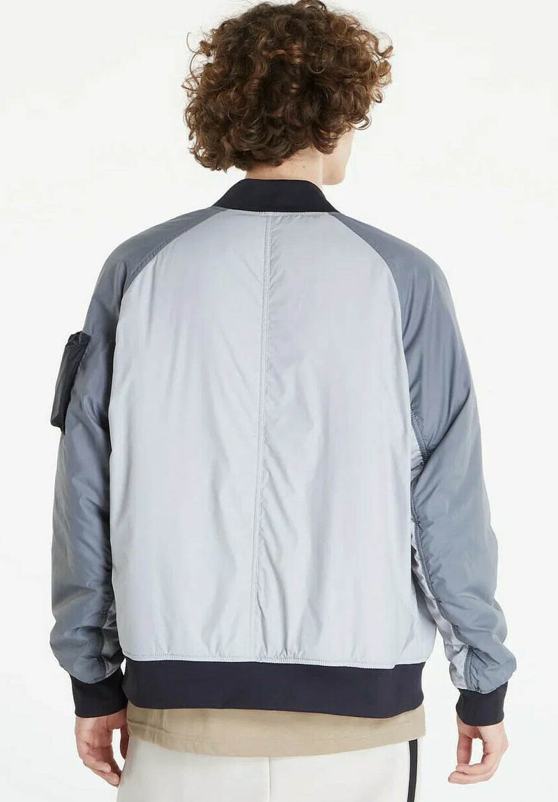 Persona con capelli ricci vista da dietro che indossa una giacca bomber grigio chiaro e grigio scuro con tasche sulla manica.