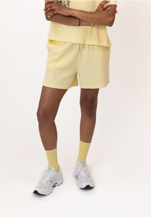 Personne portant un short jaune clair et un haut assorti, des chaussettes jaunes et des chaussures de sport blanches, debout les bras croisés.