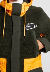 Trevlig grön fleecejacka med orange detaljer, vertikal dragkedja och framfickor. Har en vit logotyp på bröstet. Texturerad och isolerad.