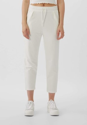 Frau trägt weiße gekürzte Jogginghosen mit elastischem Bund und Kordelzug, kombiniert mit weißen Turnschuhen vor einfachem Hintergrund.