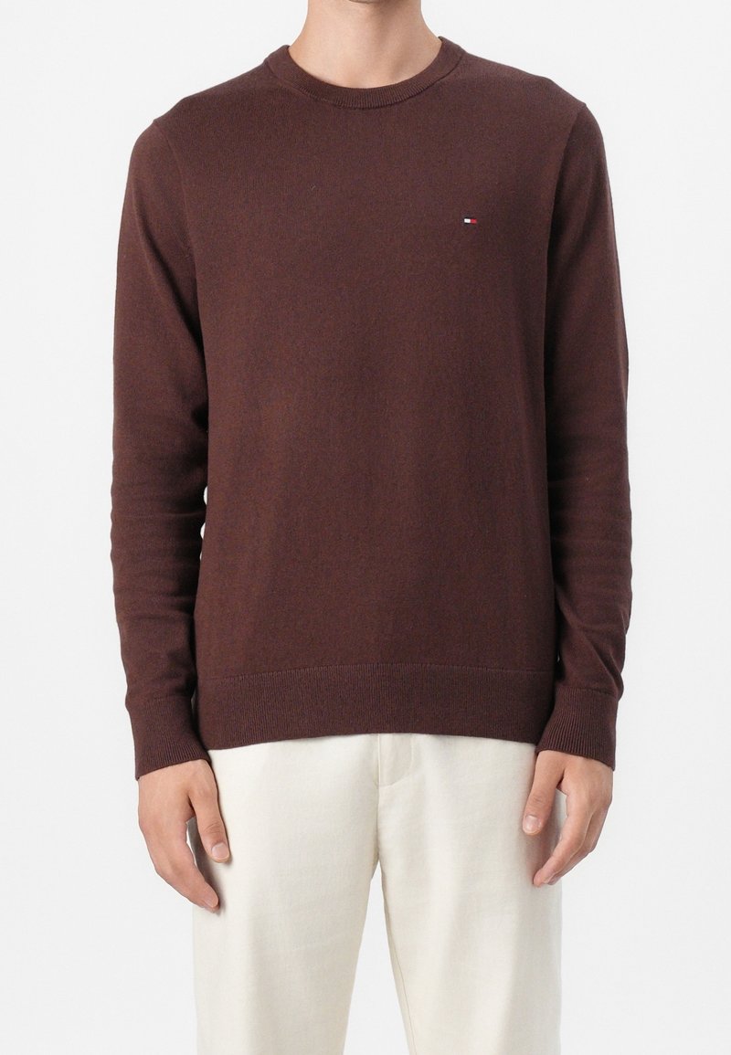 Homme portant un pull à manches longues marron avec un petit logo sur la poitrine et un pantalon crème, debout devant un fond blanc.