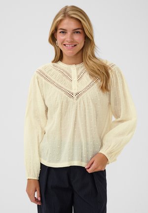 Femme souriante, portant un chemisier texturé crème à manches longues avec des détails en dentelle et un pantalon noir à fines rayures, debout devant un fond uni.