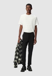 T-shirt a righe bianche, pantaloni neri e scarpe nere. Tiene in mano una camicia a quadri. Design semplice con vestibilità rilassata e forma dritta.