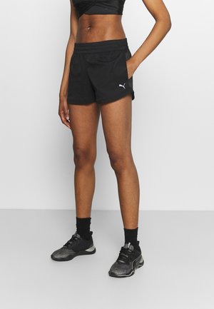 Atlet iført sorte Puma sportsshorts, sort sports-bh, sorte sokker og sorte Puma løbesko, stående mod en ensfarvet lys baggrund.