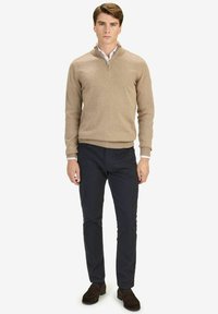 Beige Zip-Hals-Pullover mit gerippten Manschetten, kombiniert mit marineblauen Hosen und dunkelbraunen Slippern. Der Pullover hat eine glatte Textur und eine figurbetonte Passform.