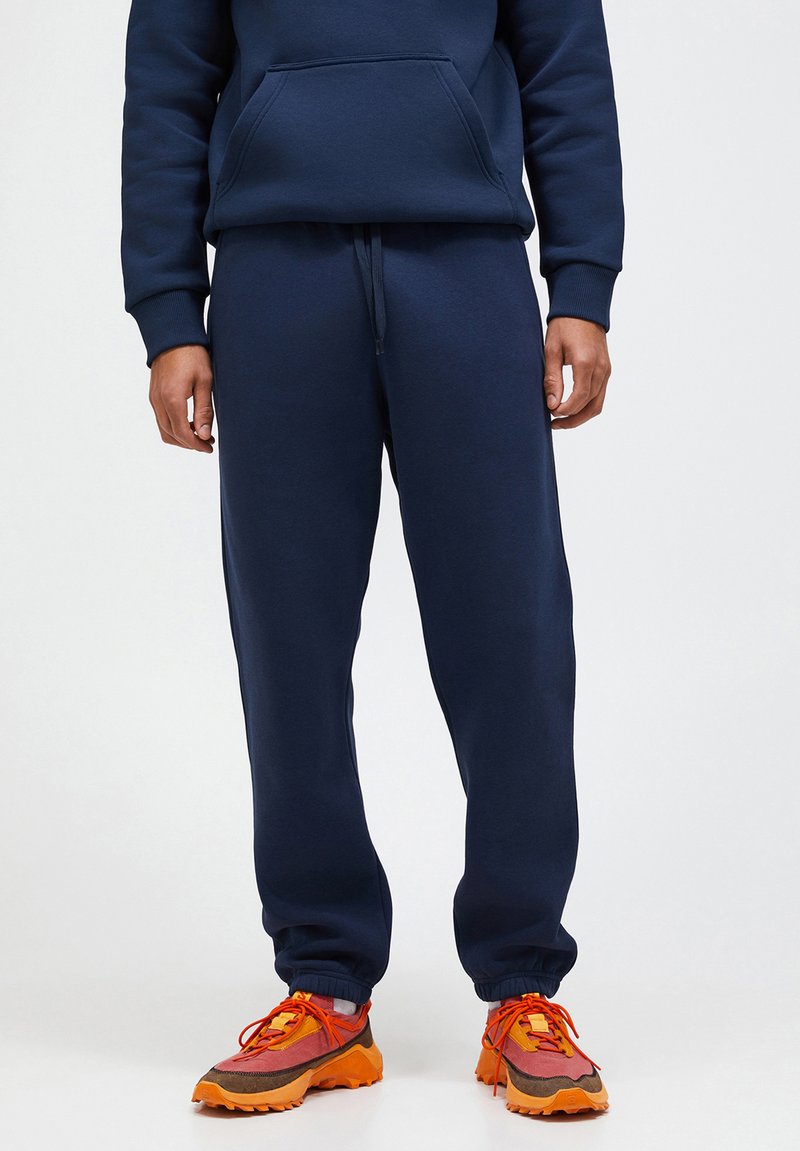 Navyblauwe joggingsbroek met elastische boorden, trekkoord in de taille, gecombineerd met meerkleurige sneakers met gedurfde oranje accenten en textielstof.