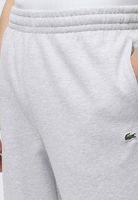 Pantalon de jogging gris clair avec taille élastique et poche latérale, avec un petit logo crocodile vert près de la poche.