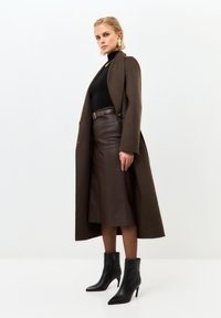 Lungo cappotto marrone in lana, abbinato a un dolcevita nero e una gonna di pelle marrone. Stivaletti neri a punta completano l'outfit.