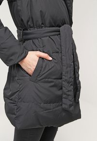 Musta, toppatakki, jossa on vyötäröllä vyö ja sivutaskut. Kankaassa on sileä tekstuuri ommelluilla yksityiskohdilla ja väljä istuvuus.