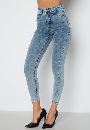 Light blue high-waisted skinny jeans made of denim, featuring front pockets, back pockets, and a subtle faded texture. Paired with black heels.