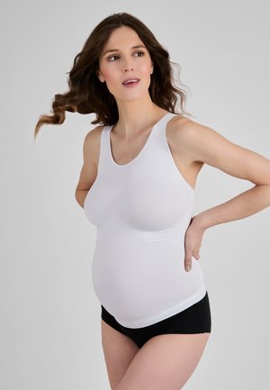 Tehotná žena s dlhými hnedými vlasmi nosí biele tielko a čierne spodné prádlo, stojí s jednou rukou na dolnej časti chrbta.