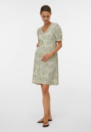 Gravid kvinde med håret sat tilbage, iført en mønstret knælængde kjole og sorte sandaler, der blidt holder om sin mave.