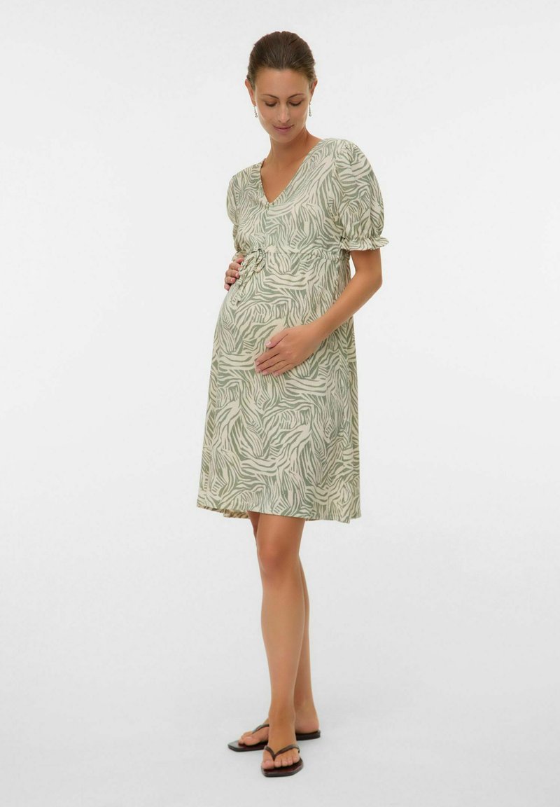 Femme enceinte aux cheveux attachés, portant une robe mi-longue à motifs et des sandales noires, caressant doucement son ventre.