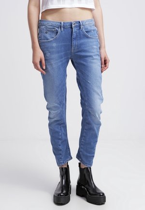 Femme portant un jean slim bleu clair légèrement usé et des bottines à plateforme noires épaisses, debout devant un fond uni.