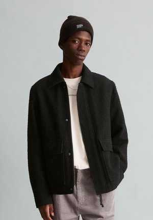 Young man wearing a black wool jacket, beige shirt, brown knit beanie, and light gray corduroy pants against a plain gray background.