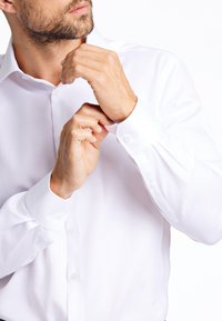 Camicia bianca a maniche lunghe con polsini abbottonati, tessuto liscio, leggera lucentezza e colletto classico, mentre viene aggiustata a mano.