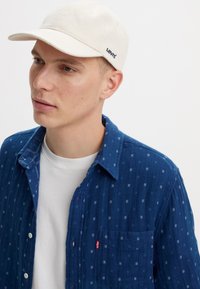 Cappello in cotone beige con visiera curva e logo blu "Levi's"; il modello indossa una camicia a quadri blu sopra una t-shirt bianca.