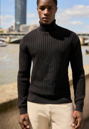 Maglione nero lavorato a maglia con texture a coste, collo alto e maniche lunghe, abbinato a pantaloni chiari in un contesto informale all'aperto.