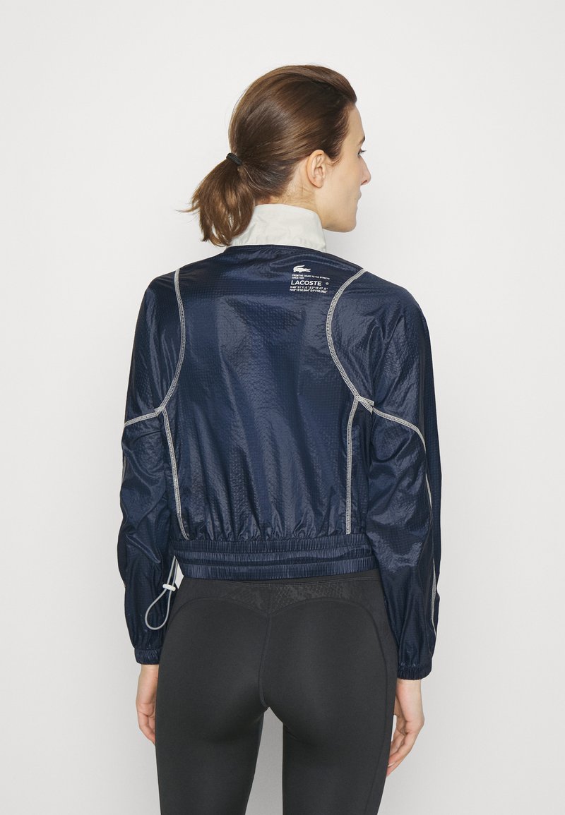 Veste légère bleu marine avec une finition texturée, taille élastique et accents réfléchissants ; dispose d'un col montant et d'une fermeture éclair.