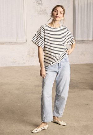 Žena stojaca v interiéri, oblečená v bielej a čiernej pruhovanej tričku s krátkym rukávom, svetlomodré džínsy a béžové balerínky s čiernymi škvrnami.