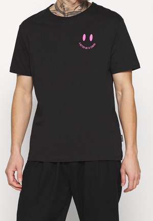 T-shirt en coton noir avec un col rond et des manches courtes, ornée d'un graphique de visage souriant rose et du texte "UN MOI MEILLEUR ARRIVE."