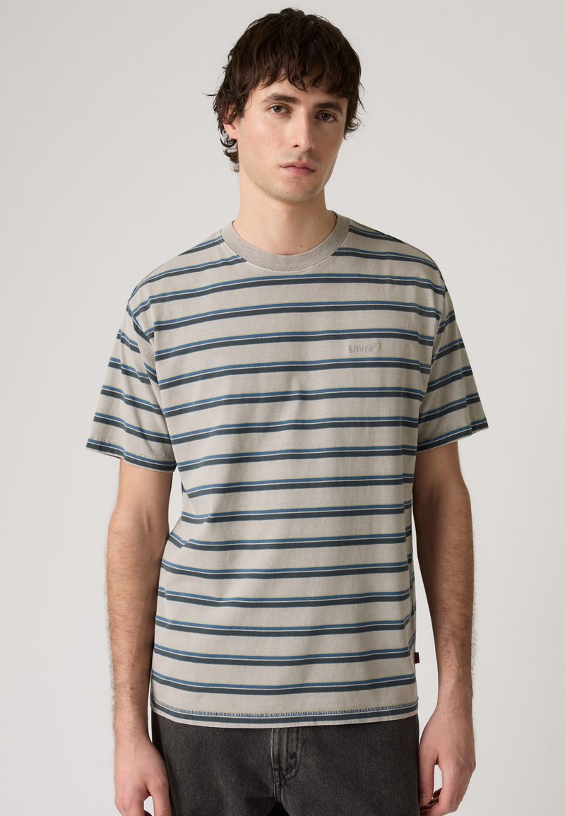 Young man wearing a beige and blue striped short-sleeve t-shirt standing against a plain light background.