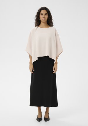 Kvinde med krøllet hår iført løs lys rosa top, lang sort nederdel, sorte spidse sko, stående mod hvid baggrund.