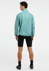 Lengvas, mėtų žalumo striukė su aukštu apykakle, elastingomis rankovėmis ir sraigteliu dizainu, derinama su juodomis dviračių šortais ir batais.