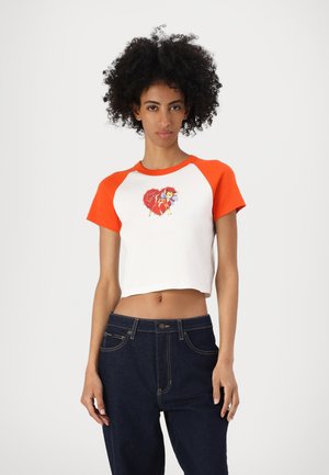 White crop top with orange raglan sleeves, featuring a colorful graphic of a character with an umbrella on the front. Dark blue denim pants.