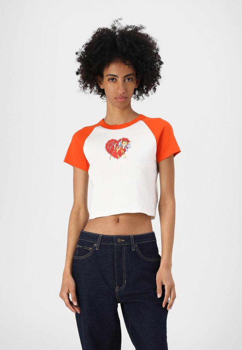 White crop top with orange raglan sleeves, featuring a colorful graphic of a character with an umbrella on the front. Dark blue denim pants.