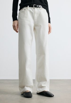 Osoba oblečená v krémových širokých džínsach s čiernym opaskom, čiernom top s dlhým rukávom a čiernymi plochými topánkami stojí na sivom podlaží.