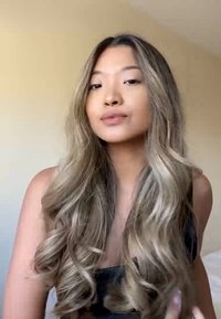 Long, wavy hair in light brown and blonde hues, wearing a black sleeveless top. Soft lighting highlights hair texture against a neutral background.