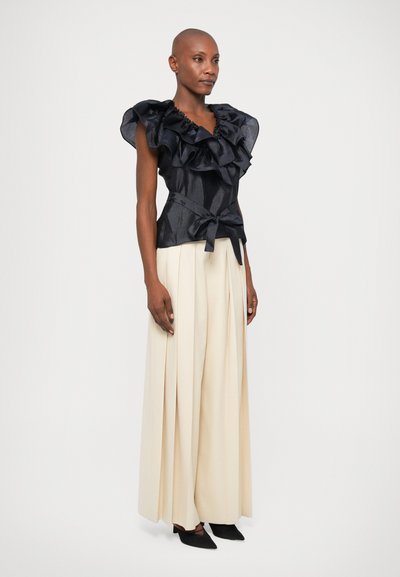 Sort flæse-top med en struktureret design og knyt-talje, parret med beige plisserede bukser og sorte spidse hæle.