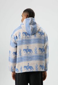 Capuchonjack in beige stof met blauwe paarden- en cactussenpatronen, met een losse pasvorm en een gestructureerd oppervlak.