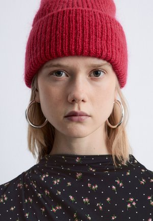 Red knitted beanie with ribbed texture, worn with a black floral-patterned shirt. Silver hoop earrings accentuate the face. Light hair is visible.