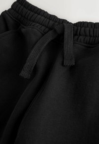 Des joggers en coton noir présentent une taille élastique avec un cordon de serrage, une texture lisse et une coupe décontractée, vus en perspective de haut.