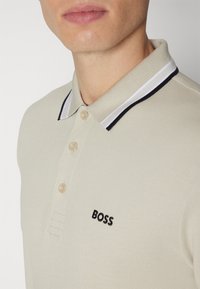 Polo beige en tissu texturé, à trois boutons, avec un col rayé blanc et marine contrastant. Il présente un logo "BOSS" noir sur la poitrine.