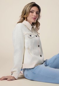 Giacca bianca strutturata con un colletto rosa chiaro a contrasto, dotata di due tasche frontali e bottoni neri, indossata sopra jeans azzurri.