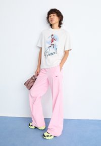 Person som har på seg en hvit t-skjorte med grafikk av en basketballspiller og teksten "Ciao!", løse rosa Nike-bukser, neonlys sneakers, og holder en liten veske.