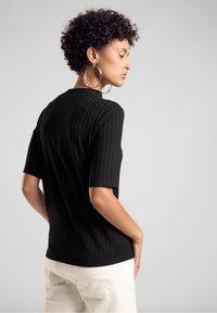 T-shirt noir à côtes avec des manches courtes, présentant un col montant et une silhouette ajustée, associé à un pantalon de couleur claire.