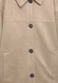 Manteau beige avec quatre boutons noirs, deux poches fendues verticales et un large col en tissu doux.
