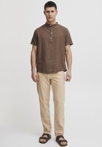 Kortærmet brun linned skjorte med opretstående krave og fem knapper, parret med lysebeige bukser og mørke sandaler.