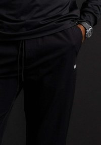 Zwarte sportbroek met een trekkoord in de taille, voorzien van een slim fit, een gladde textuur en een klein wit logo aan de zijkant.