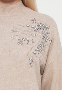 Maglione lavorato a maglia beige con decorazioni in perline argentate e cristalli a fantasia floreale sulla spalla sinistra. Tessuto morbido, collo alto.