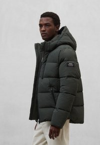 Dunjakke i mørkegrøn med hætte, lynlåslommer og et mærke på venstre ærme. Quiltet design for isolering.